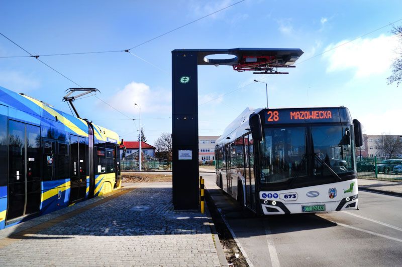 Bialy autobus miejski stojący pod ładowarką pantografową, obok niebiesko-żółty tramwaj